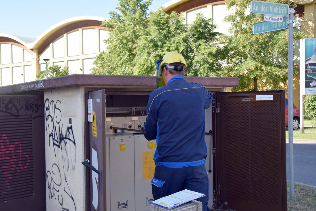 Mitarbeiter der Stadtwerke Bernau in Schutzausrüstung schaltet an der geöffneten Trafostation den Mittelspannungsleistungsschalter. Im Hintergrund ist eine Starßenkreuzung zu sehen.