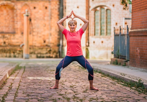 Eine Frau in sportlicher Kleidung macht eine Yogaübung auf einem gepflasterten Weg vor einer historischer Kirche.