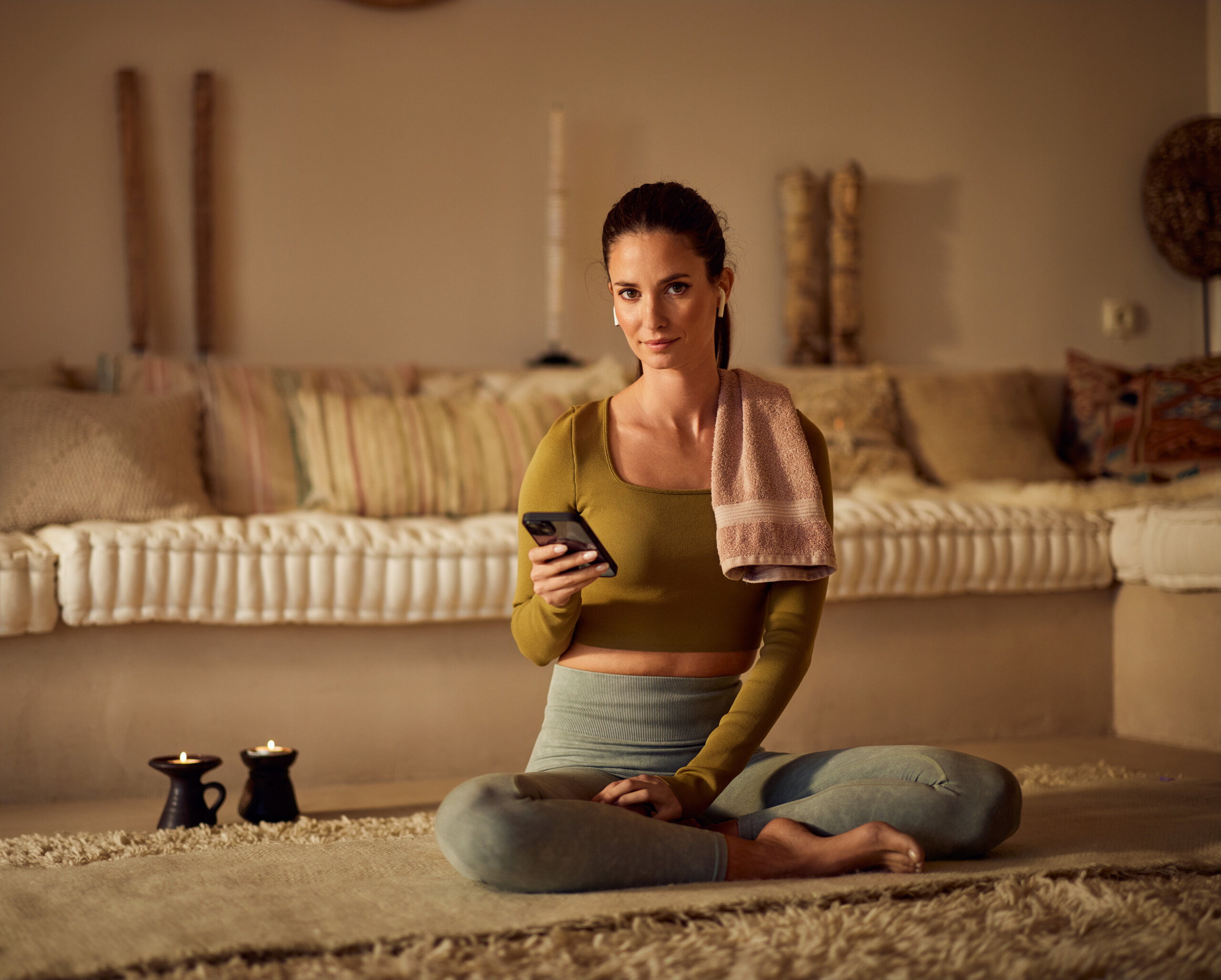 Eine Frau in Sportkleidung sitzt auf einer Yogamatte in einem gemütlich beleuchteten Wohnzimmer. Sie hält ein Smartphone in der Hand, ein Handtuch liegt über ihrer Schulter.
