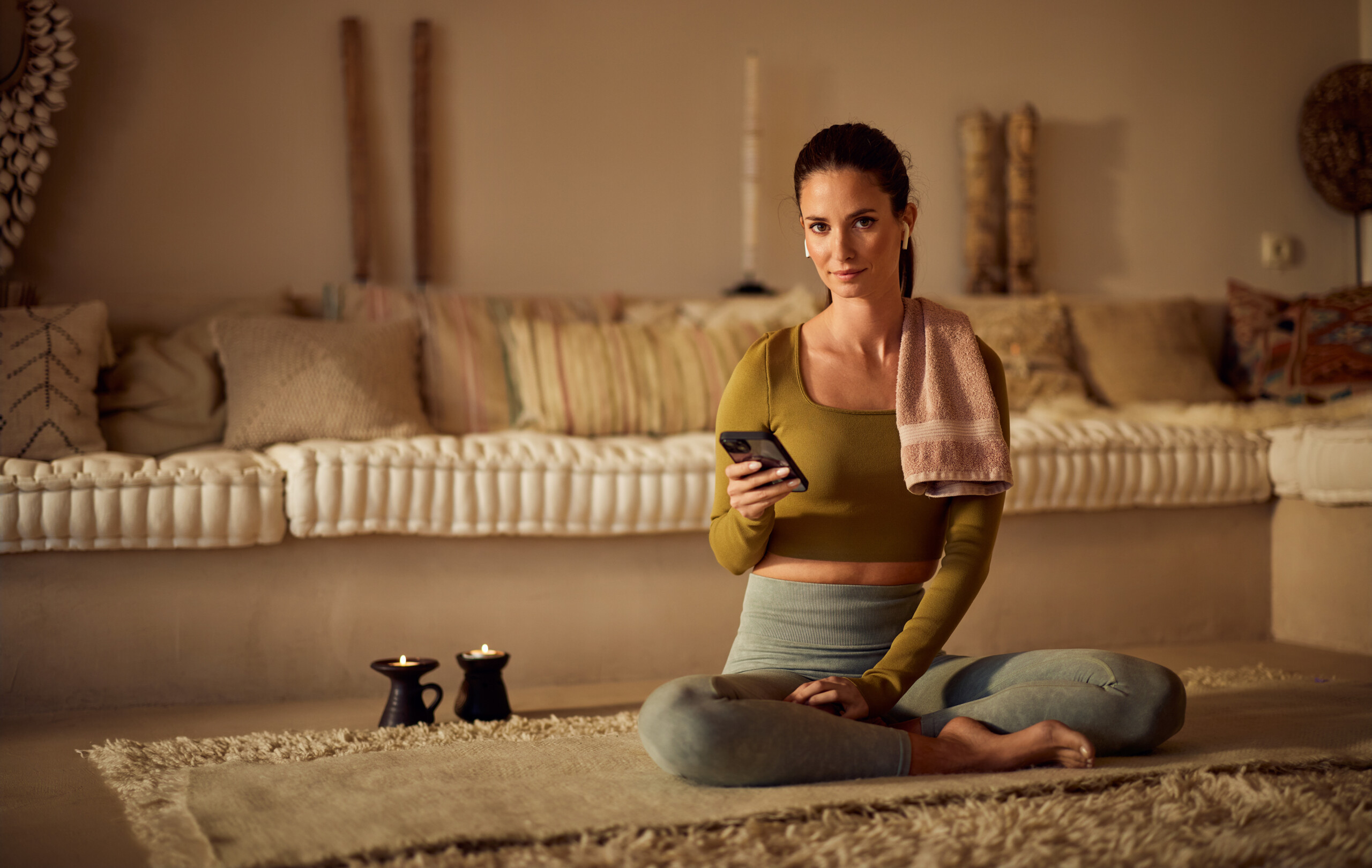 Eine Frau in Sportkleidung sitzt auf einer Yogamatte in einem gemütlich beleuchteten Wohnzimmer. Sie hält ein Smartphone in der Hand, ein Handtuch liegt über ihrer Schulter.