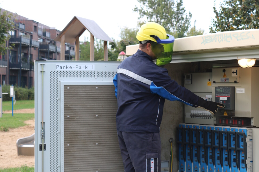 Mitarbeiter der Stadtwerke Bernau in Schutzausrüstung bedient an der geöffneten Trafostation den Niederspannungsleistungsschalter. Im Hintergrund ist ein Wohngebiet mit Spielplatz zu sehen.