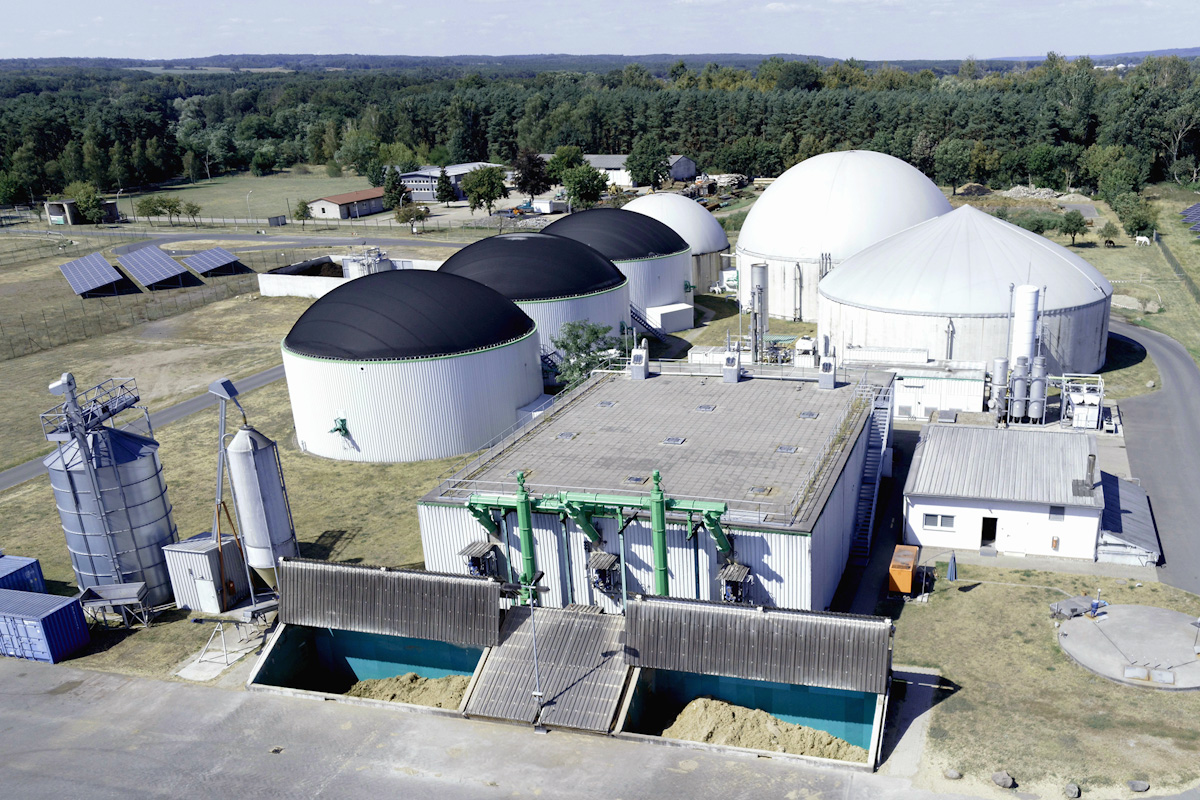 Luftbild einer Biogasanlage mit mehreren zylindrischen Garbehältern und Lagertanks umgeben von landwirtschaftlichen Feldern und Solarpaneelen.